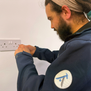 An Age Care Maintenance handyman fixing an electrical socket, wearing a navy jacket with the company’s hammer-and-nail logo on the sleeve.